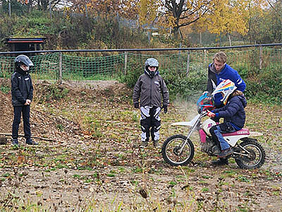 Ein Kind auf einem kleinen Motorrad wird von einem Betreuer auf einer Übungsstrecke angeleitet, während zwei weitere Kinder zuschauen.
