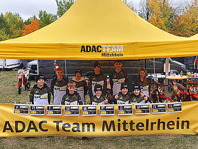 Eine Gruppe junger Motorradsportler posiert unter einem gelben Zelt mit der Aufschrift „ADAC Team Mittelrhein“. Sie halten Zertifikate und Trophäen in den Händen. Im Hintergrund sind Bäume in herbstlichen Farben zu sehen.