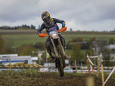 Ein Enduro-Fahrer springt mit seinem Motorrad über eine schlammige Strecke, während Dreck aufgewirbelt wird. Im Hintergrund ist eine hügelige Landschaft zu sehen.