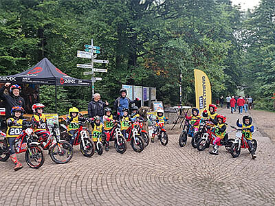 Eine Gruppe von Kindern in Motorradkleidung und Helmen sitzt auf kleinen Motorrädern. Im Hintergrund sind Zelte und Wegweiser zu sehen, während Erwachsene die Kinder beaufsichtigen.