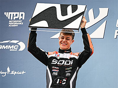 Maximilian Schleimer in Sodi-Rennanzug hält eine Trophäe hoch und lächelt bei einer Siegerehrung.