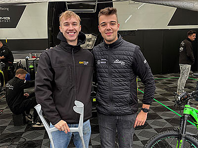 Milan Bondzio und Cedric Fuchs lächeln in die Kamera, umgeben von Rennsport-Equipment in einer Werkstatt. Milan hält Krücken, beide tragen sportliche Kleidung mit Sponsorenlogos.